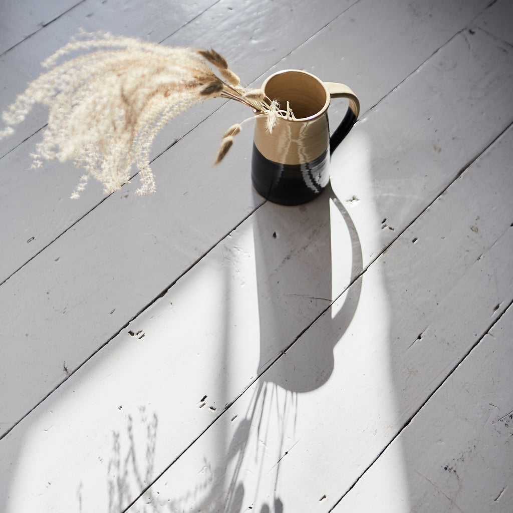 A black and beige jug containing dried pampas on a white floor