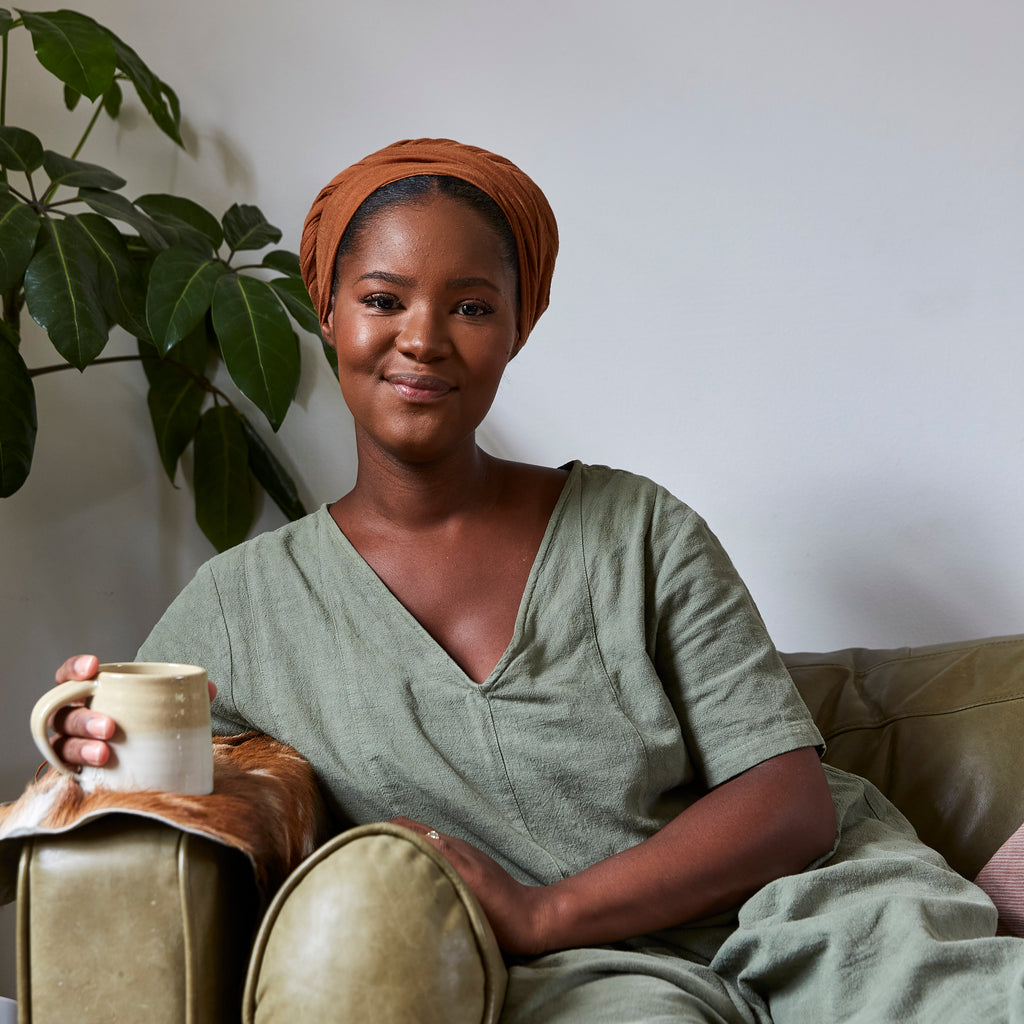 A woman with an orange head scarf and green jumpsuit sits on an olive sofa holding a mug